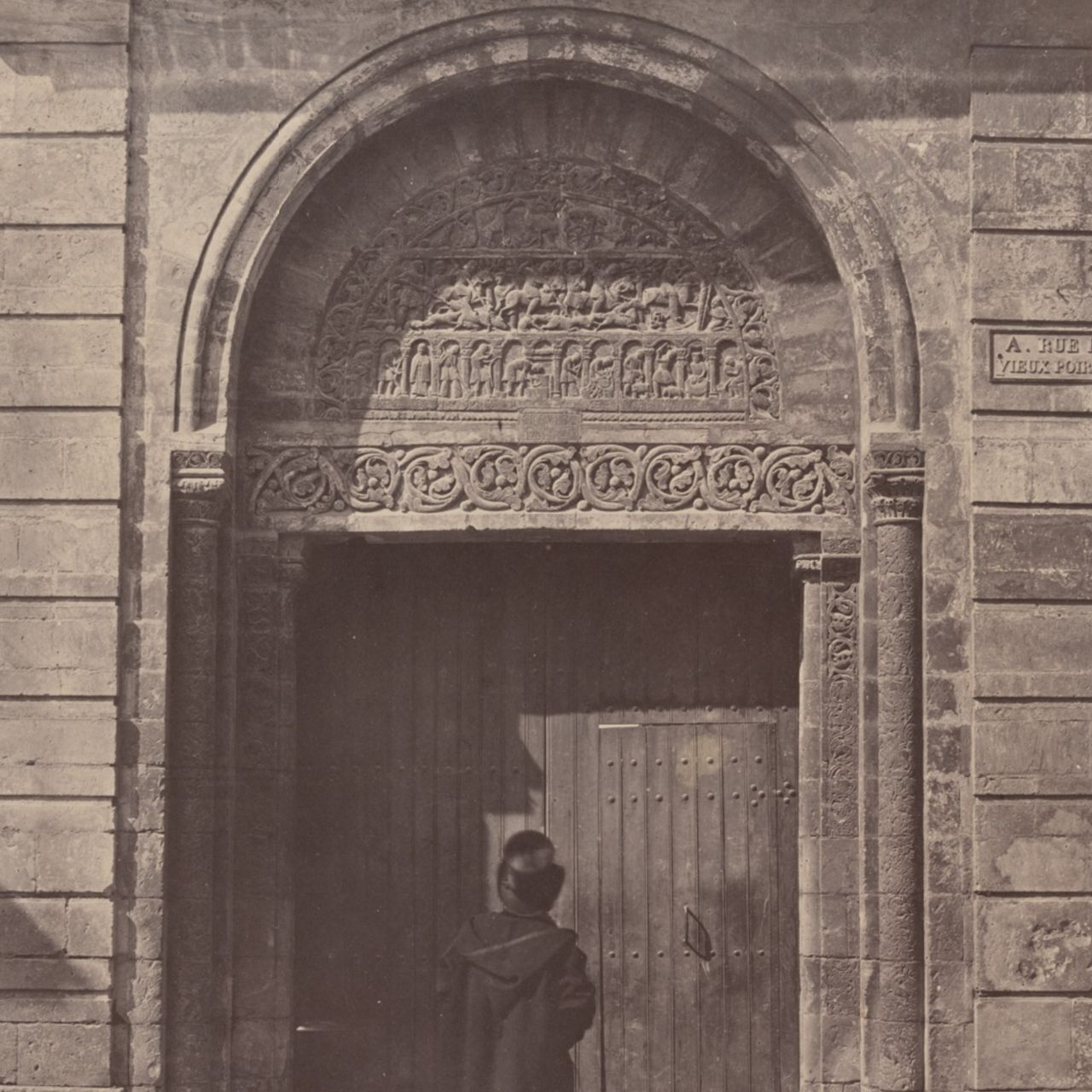 An old photograph of a the back of a man in an top-hat standing outside of a building's heavily elaborate arched stone masonry doorwaym, looking up.